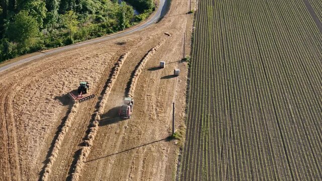 Two tractors work across a harvested field, with one forming windrows using a rake and another baling the cut crop while completed wrapped bales lie nearby, during a drone tracking flight.