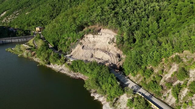 Lake Mignano and the SP21 roadway frame a major exposed landslide scar, showing slope failure, displaced sediment and erosion within the wooded Arda Valley during a drone orbit.