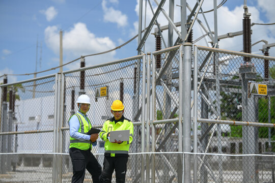 Electrical engineer inspecting electrical substation equipment with tablet beside safety fence under cloudy sky