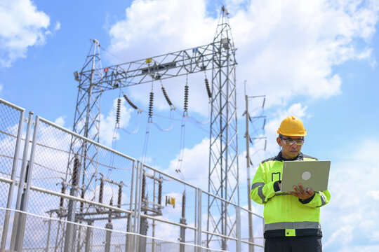 Electrical engineer inspecting electrical substation equipment beside safety fence under bright blue sky, focused on reviewing technical data and maintaining reliable power infrastructure