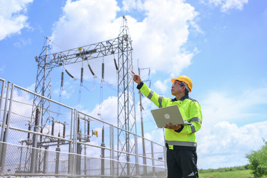 Electrical engineer inspecting electrical substation equipment outdoors beneath bright blue sky and cloud, using laptop for technical assessment and safe power system maintenance