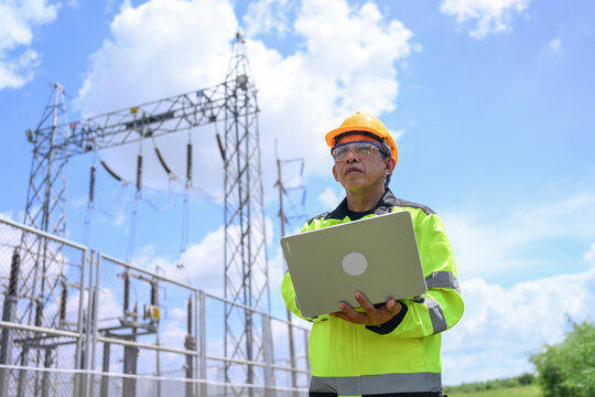 Electrical engineer inspecting substation equipment with laptop beneath bright cloudy sky, performing careful safety assessment in protective workwear