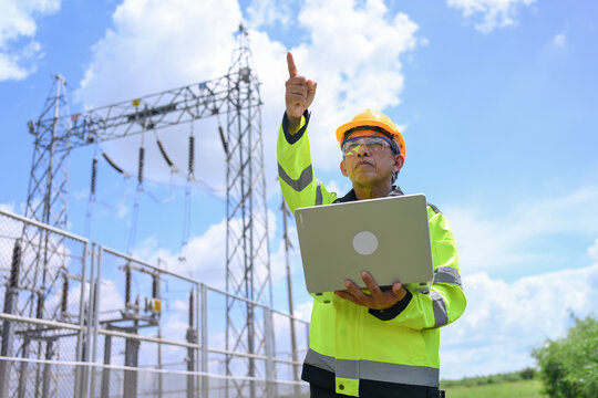 Electrical engineer inspecting power substation equipment outdoors while using laptop and pointing toward transmission structure under blue sky
