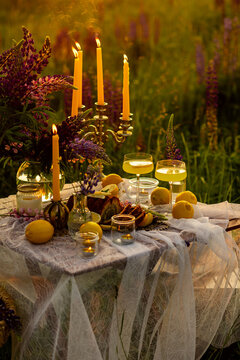 Simple countryside beautiful wedding decor, surprise romantic date outdoors, garden arrangement with lupins flowers on a meadow. Fruits, rose wine, burning candles. Sunset, candlelight, golden hour.