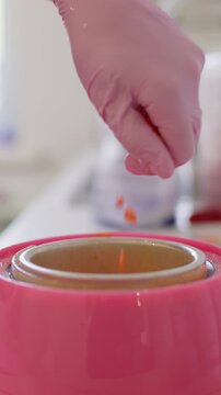 Vertical video. Wax grains. Skin care. Unrecognizable female hand in pink latex glove using orange product granules for melting in heater preparation for professional cosmetic procedure.