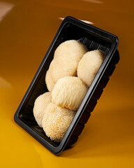 Top view of fresh Lion's Mane mushrooms arranged inside a black tray, showing varied shapes, creamy white texture and distinctive spines against a warm orange background. © DEWINs