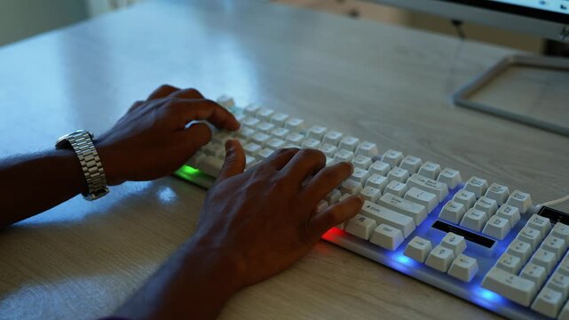 Hands typing on glowing keyboard, Close-up of hands typing on a white computer keyboard with colorful glowing lights on a desk in a dark room.