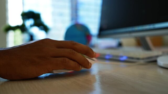 Hand using computer mouse at home office, Close-up of a hand using a computer mouse on a wooden desk in a bright home office, with a monitor and soft background blur.