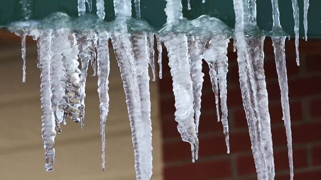 Close-up of icicles hanging from a green roof edge with brick wall background in natural daylight