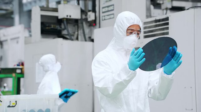 A male worker in a dustproof suit inspects silicon wafers at a semiconductor factory.