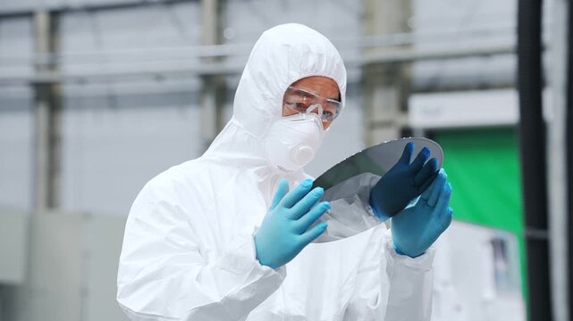 A male worker in protective clothing inspects silicon wafers at a semiconductor factory.