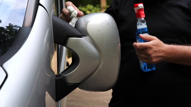 Close view of a driver cleaning the exterior side mirror using a spray cleaner during routine car care