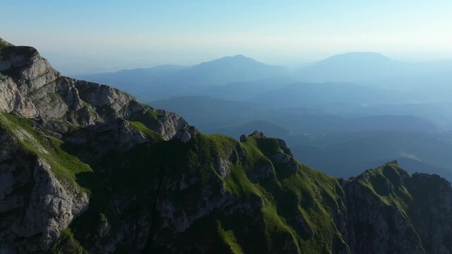 Scenic high-altitude aerial drone shot tracking along the steep green crests of the Bucegi mountains, revealing distant misty ridges in Romania.