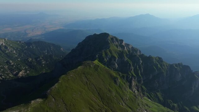 Wide aerial drone shot showcasing the rugged ridges and steep green slopes of Bucsoiu Peak in the Bucegi mountains, Romania, at daytime