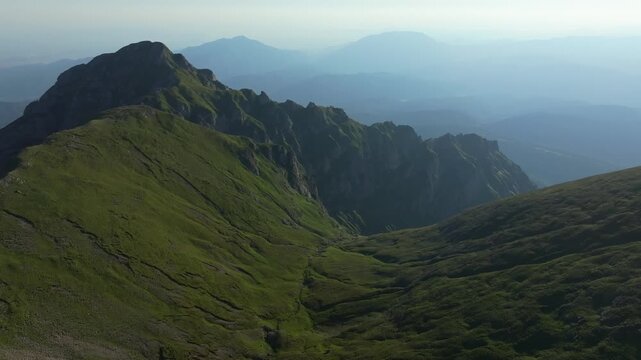Breathtaking high-altitude aerial drone shot flying over a green alpine ridge and steep rocky crests of the Bucegi mountains in Romania at daytime