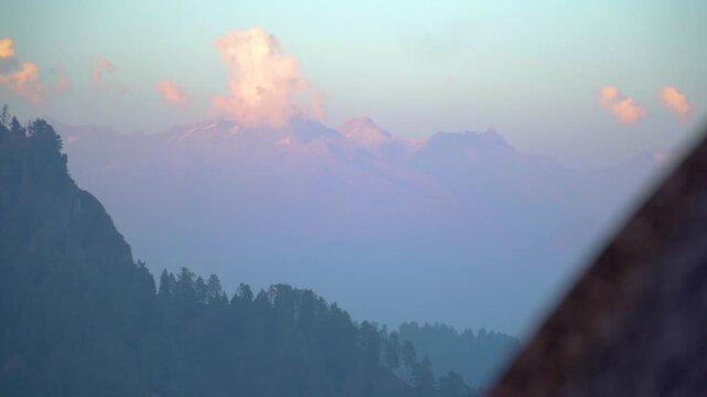 A cinematic distant shot of snow-covered Pir Panjal mountain peaks with soft sunset clouds floating above the ridge, as seen from Prashar Lake in Mandi, Himachal Pradesh, India.