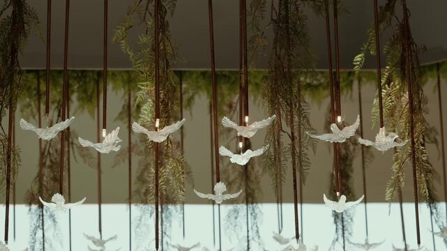 Row of glass swallow lamps on thin rods hangs among ferns and trailing greenery beneath a canopy at an event venue.