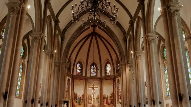 Empty church nave with pointed arches, stone columns, stained glass windows, a chandelier and an altar crucifix framed by painted panels.