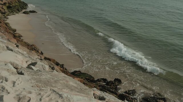 Small waves roll onto narrow sand at the foot of a rocky slope, seen looking down from above.