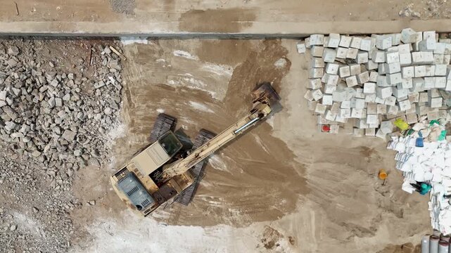Aerial top-down view of a yellow excavator moving rocks, concrete debris, and soil at active construction site. Heavy equipment performs earthwork and material handling across a prepared ground area.