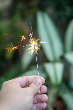Human hand holding burning sparkler with bright glowing spark and fire light bringing joyful atmosphere to celebrate special holiday outdoor in lush green nature