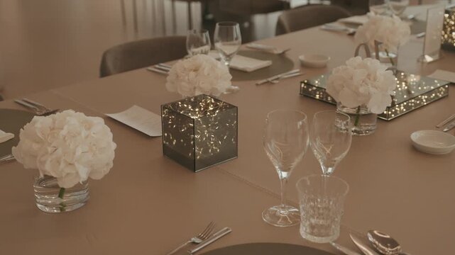 Guest table set for a wedding dinner with mirrored LED light cubes, white hydrangeas in glass vases, wine glasses and cutlery.