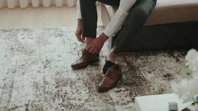 Man in green trousers and black socks sits on a bench to pull brown leather brogues onto his feet above the grey patterned rug.