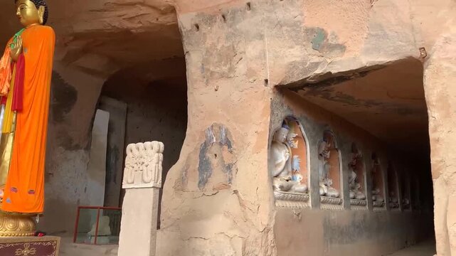 Large standing Buddha statue in orange robes inside a cave temple with rows of smaller seated Buddha niches carved into the earthen wall
