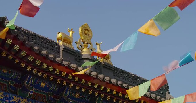 Tibetan prayer flags fluttering in wind above ornate temple roof with golden deer and Dharma wheel statues under clear blue sky