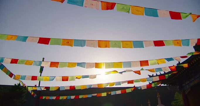 Tibetan prayer flags fluttering in wind at high-altitude temple site with sun backlighting and traditional roofline