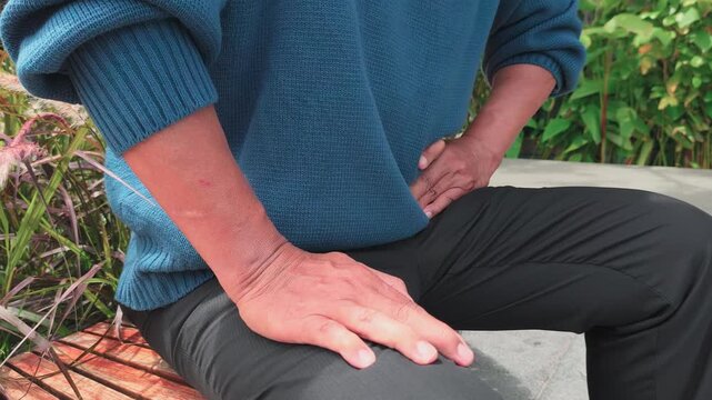 Elderly Southeast Asian man holding his left lower abdomen in pain, closeup hand on belly in outdoor park, quarter view, one hand dominant movement