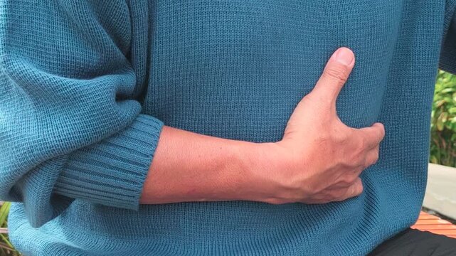 60s senior southeast asian man suffering from GERD and acid reflux at home, holding his chest with a painful expression on bench in outdoor park, hand movement closeup shot, horizontal video