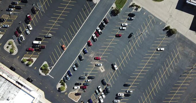 Aerial drone video of a newly paved black parking lot with yellow lines and lots of cars in front of a strip shopping mall while the drone spins counter clockwise.