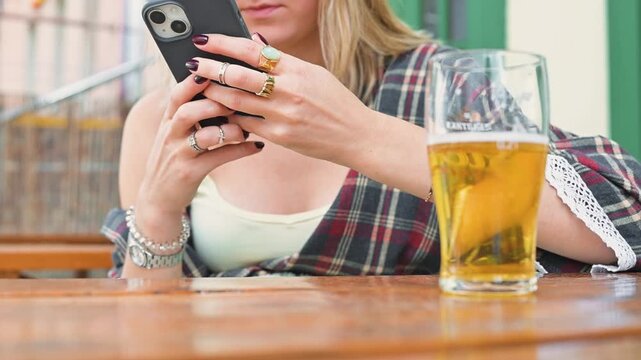 A woman with long hair is sitting at a table, focused on her smartphone while holding a glass of beer