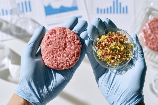 A person wearing blue gloves holds a raw plant-based burger patty in one hand and a petri dish with sprouted seeds in the other, with research charts in the background.