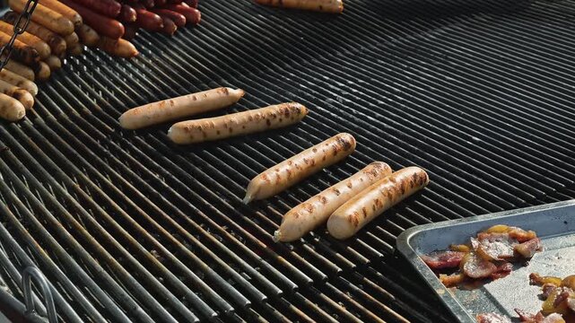 Sausages are cooking on an outdoor barbecue grill as smoke rises from the heat. A person wearing blue gloves uses tongs to turn the sausages to ensure even cooking