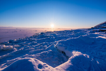 Bright winter sun shining over a frozen coastal landscape in Finland, featuring cracked sea ice, snow-covered formations, and a clear blue sky during sunset. © Artem