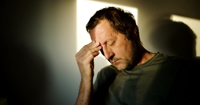 Man suffering from a severe headache, pressing his fingers to his temple in pain, dramatic window light and dark shadows indoors. 4K.