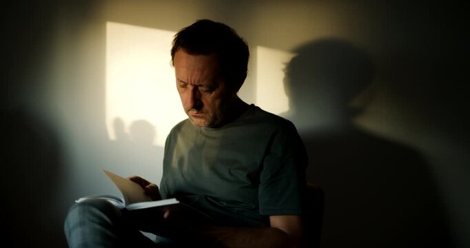 Man reading a book indoors in dramatic window light, focused expression with strong shadow and contrast on the wall. 4K.