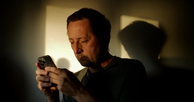 Man using his smartphone in dramatic window light, strong shadow cast on the wall, moody indoor portrait. 4K.