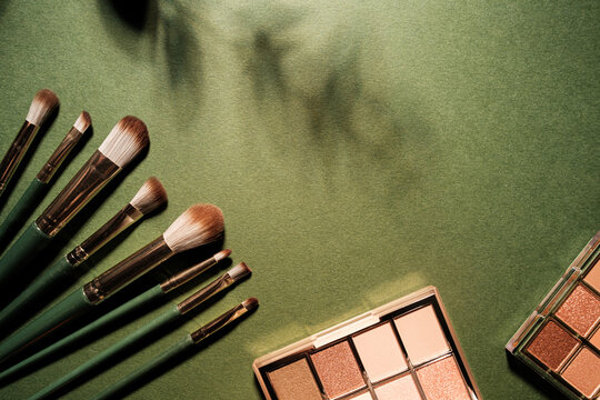 Makeup brushes and eyeshadow palettes arranged on a textured green background with shadow patterns