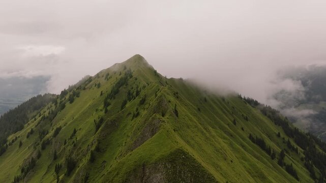 Aerial view of the narrow Arnigrat ridge in Switzerland with steep grassy slopes and scattered conifers as low clouds wrap the summit and partly conceal the surrounding alpine valleys