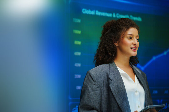 Hispanic Woman gives a business presentation in front of a digital dashboard with charts and graphs, highlighting data analytics, performance and strategic insights.