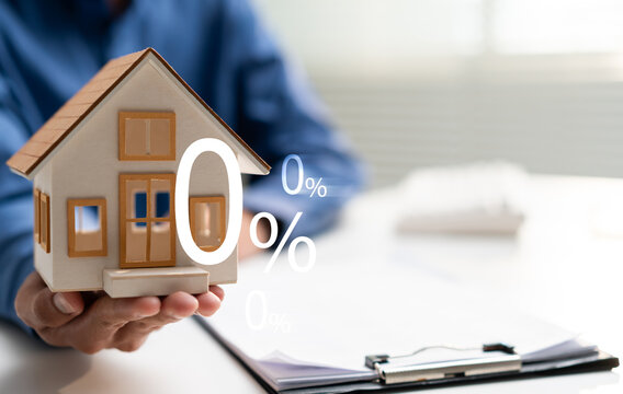Hand holds a model house with zero percent symbol beside financial documents, representing interest free home financing, mortgage promotion, property investment and affordable housing loan.