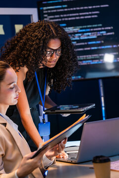 A female developer duo read through the source code on screen in a dark office wall papered with code screens, an image of women in software engineering in a modern technology workplace