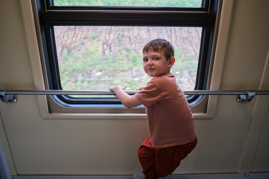 Little child looking out of a train window while traveling. Young boy standing in a passenger carriage watching the scenic landscape pass by. Tourism, vacation and family journey trip.