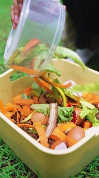Woman pouring vegetable scraps and food waste into container for composting on green lawn. Food waste recycling, zero waste, eco-friendly, and sustainability concept. Vertical Shot