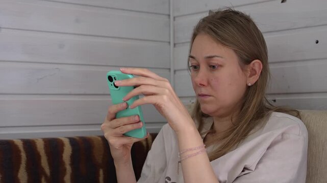 Young woman sits on a sofa, deeply absorbed in her glowing mobile device. Her gaze remains locked on the screen, reflecting a state of modern obsession and constant engagement with online worlds.
