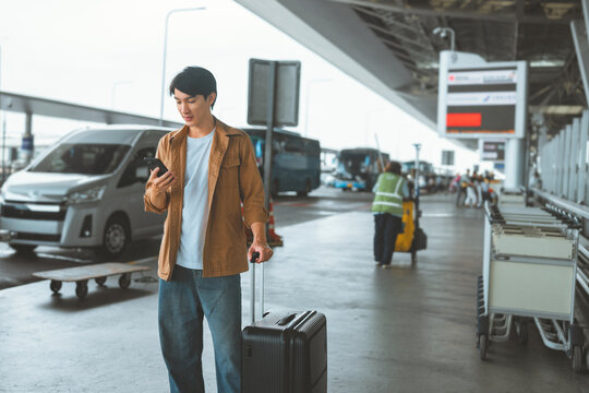 Young Asian male traveler using smartphone to check map or find airport pickup transportation outside terminal, standing with rolling suitcase near vehicle drop off area, navigation, travel planning