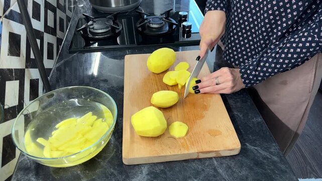 An adult cuts potato sticks on a wooden board beside a bowl of soaking potatoes, home cooking, meal preparation, knife skills.